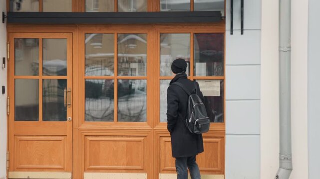 Man in black coat and beanie stands outside observing wooden doors with glass panels. Man carries backpack while background shows snow on ground near building exterior. scene is calm