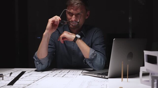 Tired man experiencing tension with hands on head, having a headache from overworking and stress, sitting at a desk with blueprints and a laptop late at night
