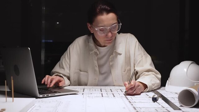 Female architect working late on detailed building blueprints using ruler and pencil, modern engineer designing construction plans at desk with laptop and hardhat