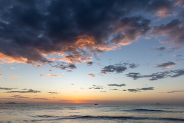 Obraz premium A lone fishing boat drifts on a calm sea as the sun sets beneath a dramatic sky, painting clouds with warm orange and purple tones and reflecting on gentle, rippling waves. Cam Ranh, Vietnam 