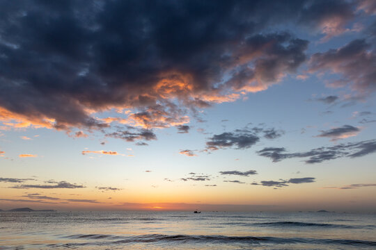 A lone fishing boat drifts on a calm sea as the sun sets beneath a dramatic sky, painting clouds with warm orange and purple tones and reflecting on gentle, rippling waves. Cam Ranh, Vietnam 
