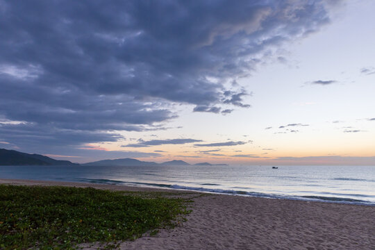  Soft pastel sunset over a tranquil beach seascape with gentle waves, low coastal vegetation and a lone boat on the horizon beneath dramatic cloud formations, Cam Ranh, Vietnam