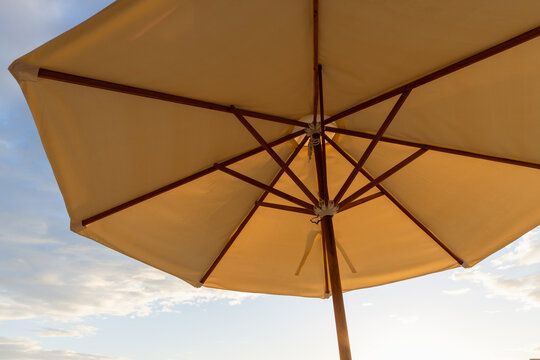 Sunlit beach umbrella photographed from below, showing wooden ribs and canvas canopy against a soft blue sky at sunset, evoking summer relaxation, seaside vacation and warm golden light