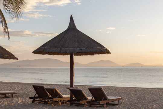 Empty wooden lounge chairs sit beneath a large thatched umbrella on a quiet sandy beach at sunset, with calm sea, pastel sky and hazy islands on the horizon creating a peaceful tropical atmosphere