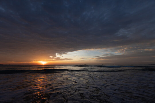 A dramatic ocean sunset lights the horizon, casting warm orange reflections across wet sand and gentle waves beneath a moody cloudscape, evoking calm, solitude and coastal beauty. Cam Ranh, Vietnam