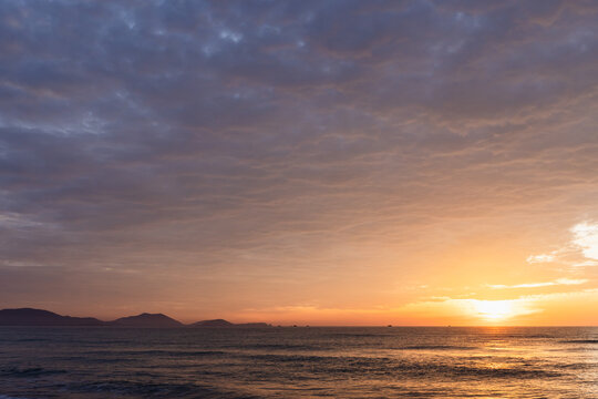 Golden sun sinks toward the horizon casting warm light across a calm ocean, silhouetting distant islands beneath a vast, dramatic cloudscape. Evokes peace, solitude and coastal tranquility at dusk