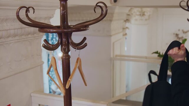 Man in dark coat hangs his black leather backpack on ornate wooden hook of brown metal stand then removes it and walks away. scene features decorative back of white architectural molding