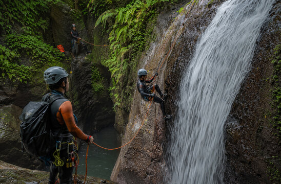 Canyoneering adventurers navigate a majestic waterfall, rappelling down a rocky cliff amidst lush, tropical foliage. A guide observes from a safe distance, ensuring a thrilling and safe expedition.