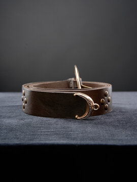 Brown leather belt with decorative crescent hook and metal studs on dark surface.