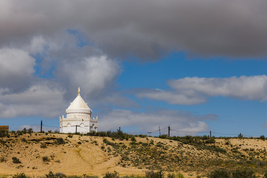 View of the white domed Karynzharyk mausoleum set against a dramatic cloudy sky in the arid desert landscape of Karakiya District, Mangystau Region, Kazakhstan.