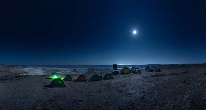 View of a campsite with tents and off-road vehicles under a bright moon and starry sky at the Bozjyra rock formations in Aktau, Mangystau Region, Kazakhstan.