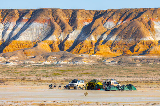 View of Gora Bokty with its colorful striped sedimentary rock formations rising above a desert campsite with tents and off-road vehicles in Karakiya District, Mangystau Region, Kazakhstan.