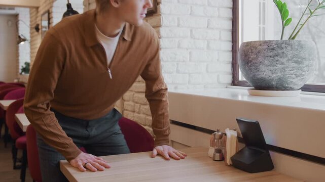 Man in brown sweater moves through and sits at empty table inside cozy cafe with red chairs wooden tables white brick walls hanging lamps framed pictures on wall large windows with potted