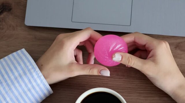 Hands stretch pink squishy stress toy at home workspace with coffee and laptop in the background. Concept of remote work, work-life balance, stress relief, relaxation and self-care during the workday.