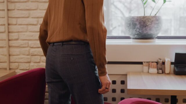 Young man in brown ribbed sweater and gray plaid pants stands moves to sit at wooden table with pink chairs. setting features indoor soft lighting brick walls window views plants