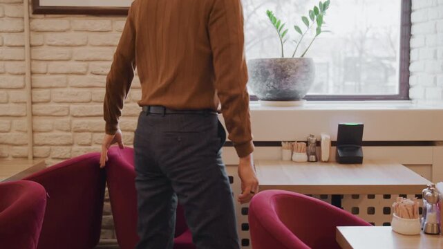 Man sits at table in indoor setting looking out window and then stands up to leave. scene features brick walls red chairs natural light from outside plants on tables condiment holders napkin