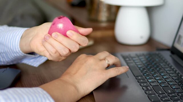 Woman squeezing one hand pink squishy stress toy while other work on laptop using touchpad in 4k footage. Concept of multitasking, work stress relief and tactile coping during digital tasks.