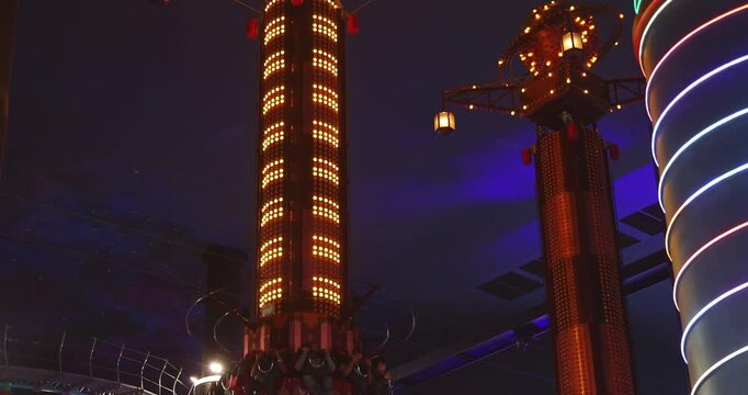 Genting Highlands, Malaysia, 24 Feb 2026. People sitting on drop tower ride at indoor amusement park. Illuminated towers with neon lights, dark background. Documentary of theme park attractions.