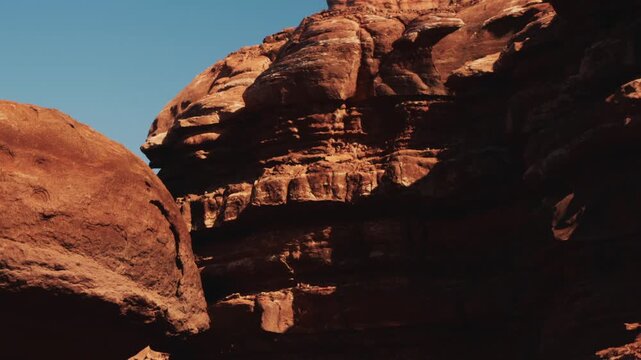 Visitors walk through a desert canyon. The tall red rock formations rise high against a clear blue sky. Sunlight casts shadows on the rugged landscape.
