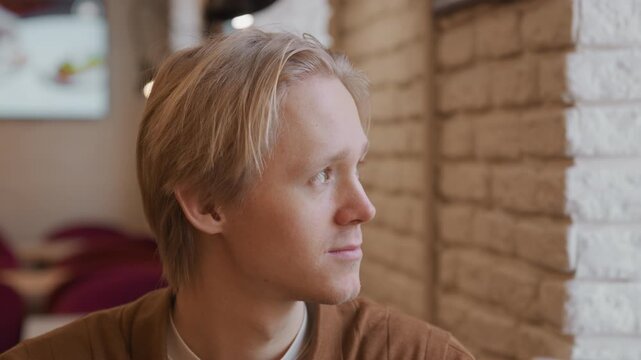Close up of young man with short blonde hair wearing brown shirt looking around in indoor setting. background features soft lighting and blurred elements like brick walls and hanging lamps