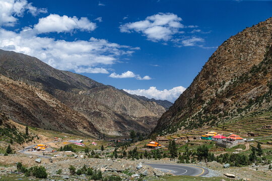 View of a mountain valley with a winding road, small houses, and terraced fields under a clear blue sky in Balakot, Khyber Pakhtunkhwa, Pakistan.