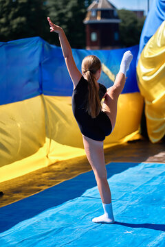 Young gymnast performing a high leg stretch on a blue mat during outdoor gymnastics practice, showing flexibility, balance and concentration in bright sunlight with colorful background banners.