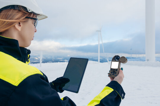 Wind energy technician checks data on tablet and anemometer in snowy landscape