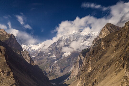 View of the rugged, snow-capped peaks of the Karakoram mountain range under a deep blue sky with drifting clouds in Hunza Nagar, Gilgit Baltistan, Pakistan.