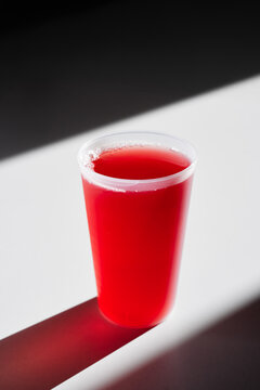 A vivid red juice fills a clear plastic cup, set on a stark diagonal shadow against white and black background. Crisp, minimal composition emphasizes color, freshness, and modern studio styling.