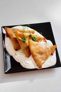 Golden fried triangular pastries rest on bed of creamy white sauce, lightly garnished with microgreens for fresh, modern presentation. for gourmet appetizers, stylish dining, and food photography.