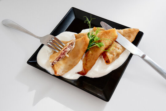 Golden fried pastry turnovers filled with meat rest on bed of creamy sauce, topped with a fresh herb garnish. A stylish plated dish with fork and knife for an elegant dining vibe.