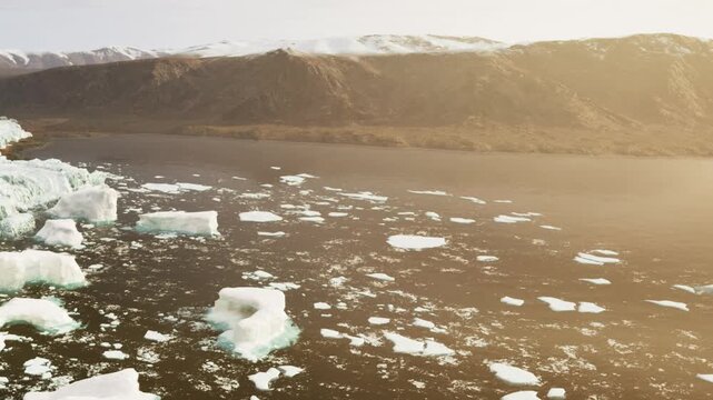 Large icebergs drift in the water close to a rocky shore under a clear sky. Mountains rise in the background as the sun sets, casting soft light on the landscape.