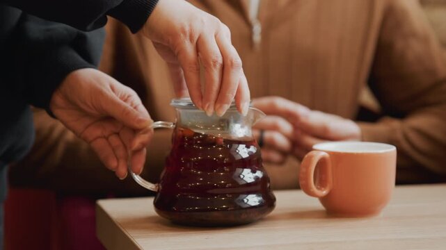Close up of hands preparing coffee with glass carafe and pink cup on wooden table. Hands pour liquid into pot then remove lid to reveal dark brew inside. Blurred background shows another