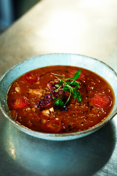 rustic ceramic bowl of hearty tomato-based seafood soup topped with a grilled octopus tentacle, cherry tomatoes and fresh herbs, presented on a metal tabletop for a warm, savory restaurant-style feel.