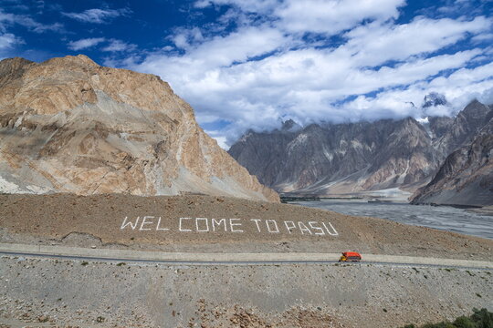 View of the Passu Cones Viewpoint with the Welcome to Pasu sign on the hillside, a truck on the highway, and rugged mountain peaks under a cloudy sky in Hunza Nagar, Gilgit Baltistan, Pakistan.