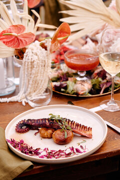 Plated grilled octopus served with roasted potatoes, microgreens and vibrant edible flowers on rustic wooden table, surrounded by wine, salad and elegant table decor for a warm fine dining experience