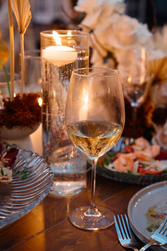 Soft candlelight reflects in a glass of white wine beside floral centerpieces and plated appetizers, creating an intimate, warm atmosphere for a romantic dinner or elegant evening celebration.