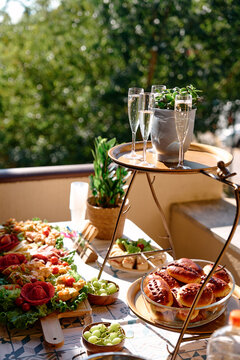 Outdoor Brunch Table With Champagne Flutes, Appetizers, Bread Rolls and Greenery on Balcony