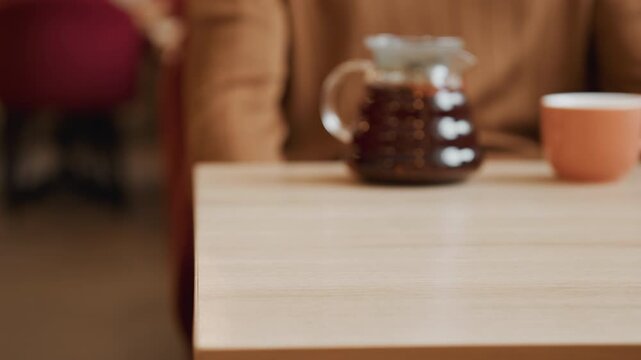 Close up of hand gently moving and placing empty woven wicker basket on light wooden table. Background includes blurred coffee carafe cup warm indoor setting with soft lighting