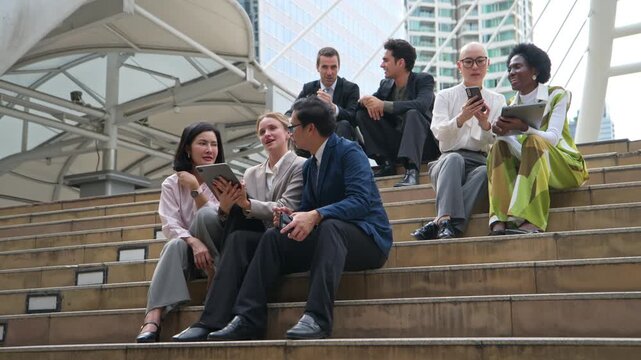 International business professionals from diverse ethnic backgrounds sitting together on city steps, discussing ideas and using digital devices in an urban setting
