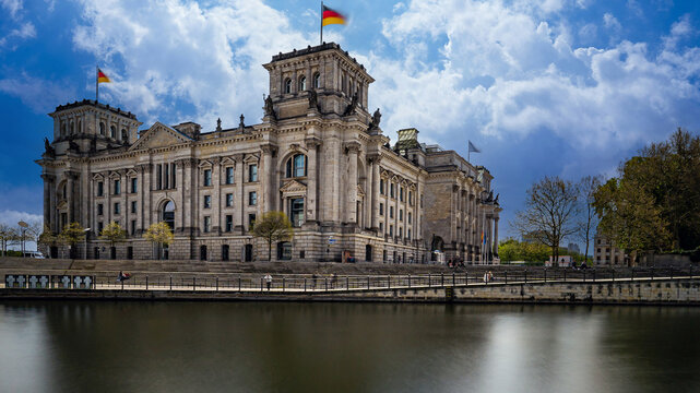 View of the Reichstag Building with its historic stone facade and German flag flying under a dramatic cloudy sky along the riverbank in Berlin, Berlin, Germany.