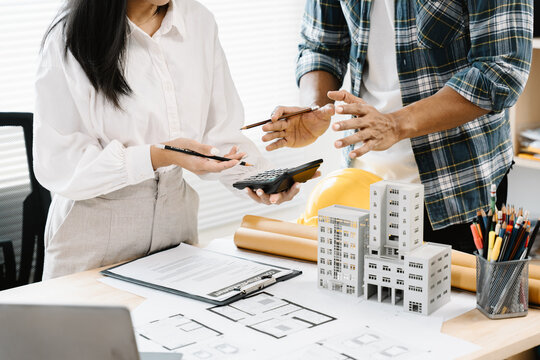 Architectural Planning Session: Two professionals meticulously review blueprints and architectural models, using a calculator and discussing plans for a building project.