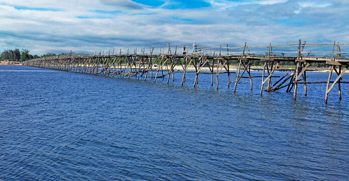 View of the rustic Ong Cop wooden bridge stretching across the water with people walking on the planks under a blue sky in Tuy An Dong, Dak Lak, Vietnam.
