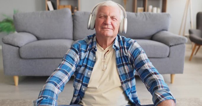 Senior man wearing headphones listening music at home. He sits near the sofa, eyes closed and smiling, enjoying wireless audio in a bright living room. Calm relaxation and leisure theme.