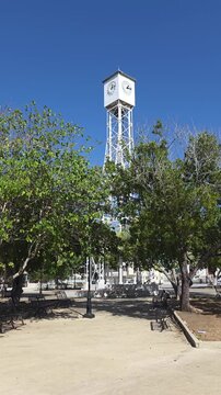 Historic Steel Clock Tower in Main Plaza framed by Green Trees against Clear Blue Sky in San Fernando de Montecristi, Dominican Republic, Vertical 4K Video