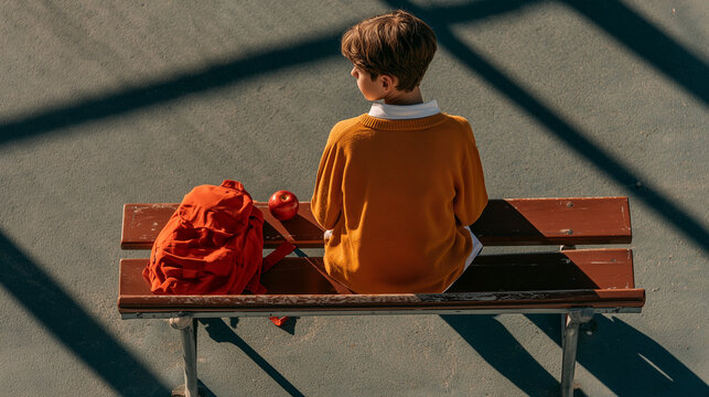 Rear view of a schoolboy sitting alone on a wooden bench with a red backpack beside him, empty playground, student waiting, school isolation, childhood loneliness, after school, elementary pupi