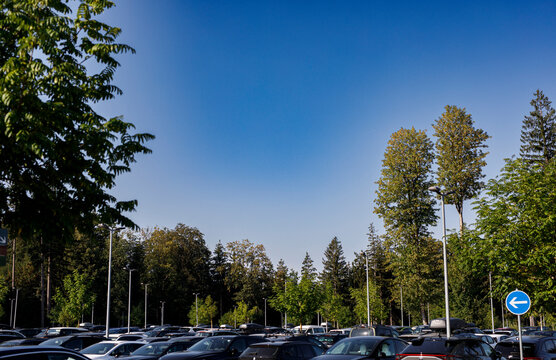 A full outdoor parking lot under a clear blue sky, surrounded by green trees and streetlights.