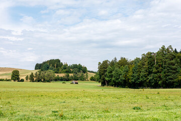 Naklejka premium Expansive green fields stretch towards rolling hills covered in dense forest under a cloudy sky, wit