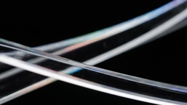 Abstract macro shot of two curved transparent glass or plastic rods crossing each other with iridescent light reflections against a dark black background