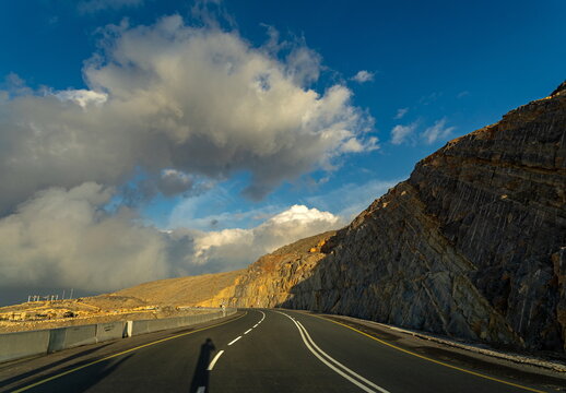 View of a winding asphalt road cutting through rugged rocky mountains under a dramatic blue sky with fluffy white clouds in Ras Al-Khaimah, Ras Al Khaimah, United Arab Emirates.
